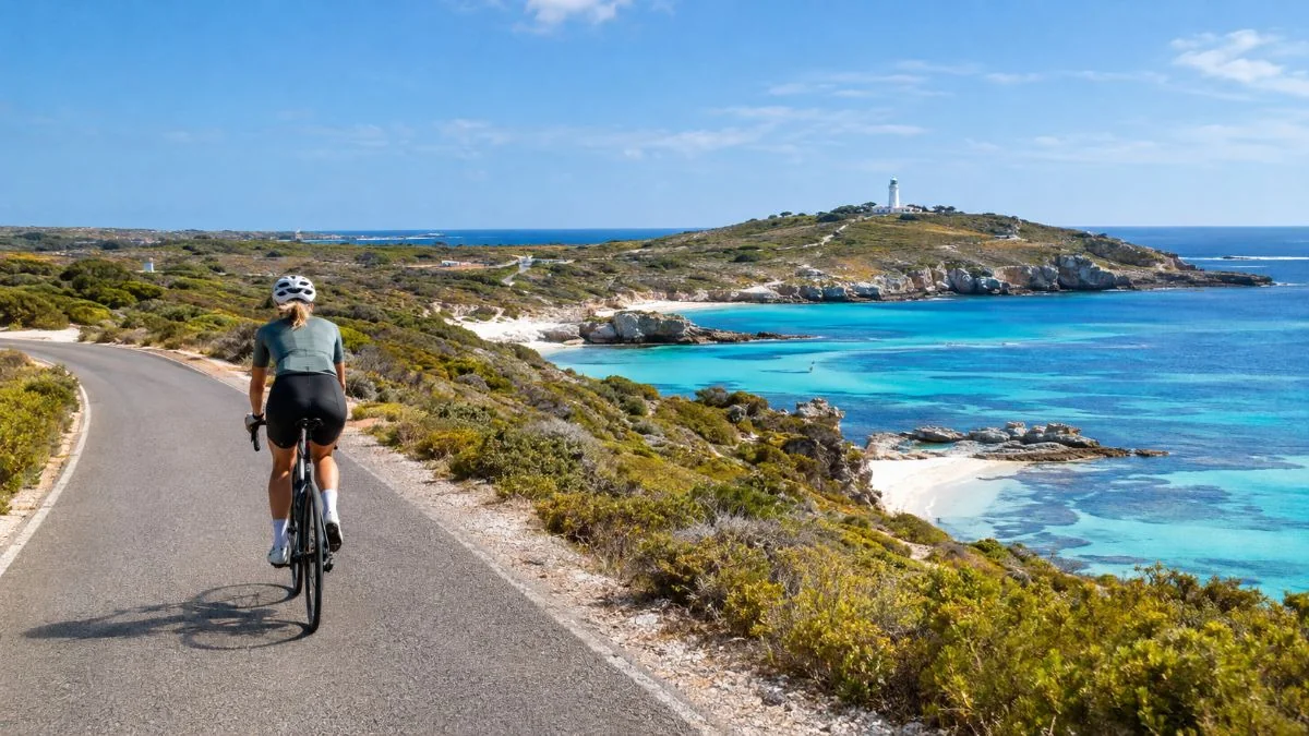 Un ciclista che pedala su una strada costiera tranquilla sopra baie turchesi con un faro su una collina lontana